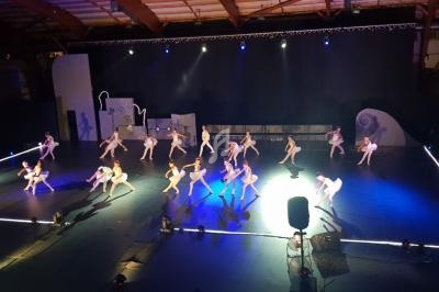 Un groupe de danseurs en tenue blanche effectue une chorégraphie sur une scène éclairée dans une grande salle.