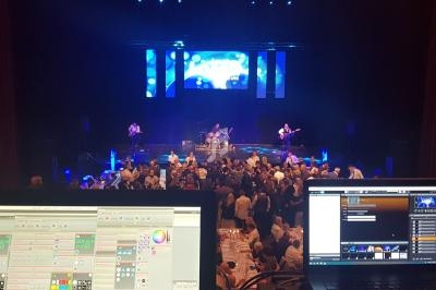 Vue depuis une régie technique sur un concert avec musiciens sur scène et public dans une salle éclairée.