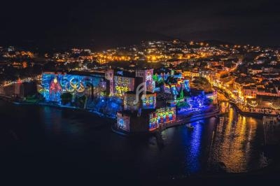 Vue nocturne d'une ville côtière illuminée avec des projections colorées sur un château et des lumières dans les rues.
