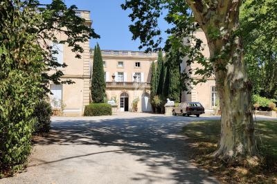 Allée ombragée menant à une grande bâtisse en pierre entourée d'arbres, avec une voiture stationnée devant.