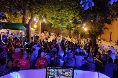 Vue d'une fête de village en soirée avec un DJ en arrière-plan, des lumières et une foule dansant sous les arbres.