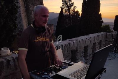 Un DJ mixe en plein air au coucher du soleil, devant un mur en pierre et des arbres.