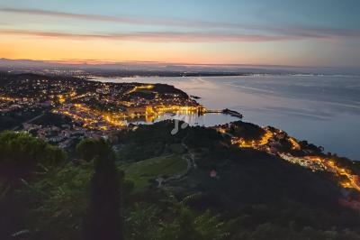 Vue nocturne d'une ville côtière illuminée avec un port, entourée de collines et bordée par la mer au coucher du soleil.