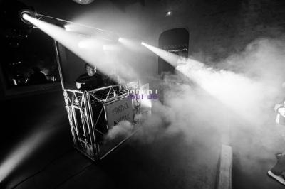 Un DJ mixe sur une scène éclairée par des projecteurs, entouré de fumée dans une ambiance nocturne.