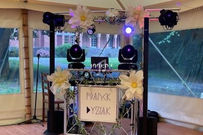 Structure de DJ décorée de fleurs blanches, éclairée par des projecteurs, installée sous une tente avec des murs…