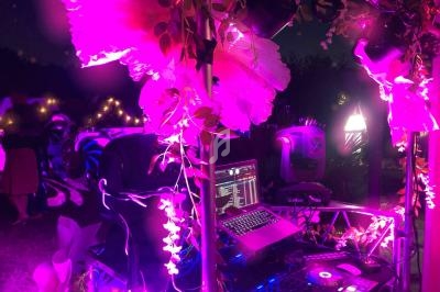Poste de DJ éclairé par des lumières roses et violettes, entouré de décorations florales dans un cadre nocturne.