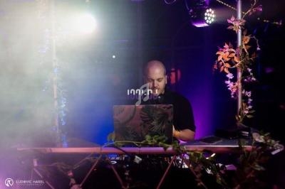 Un DJ mixe sur une scène décorée de plantes, éclairée par des lumières colorées et entourée de fumée.