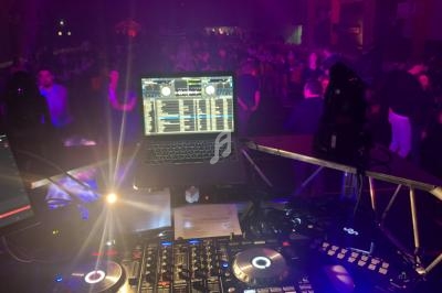 Poste de DJ avec platines, ordinateur portable et éclairage, face à une salle remplie de personnes dans une ambiance festive.