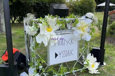 Poste de DJ décoré de grandes fleurs blanches, installé en extérieur sur une pelouse avec des arbres en arrière-plan.