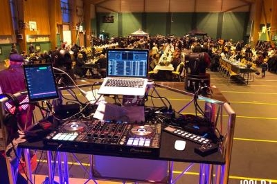 Poste de DJ avec platines et ordinateur portable, face à une salle remplie de tables et de convives dans un gymnase.