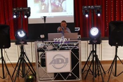 Un DJ anime un événement avec des équipements audio et lumières, devant un écran de projection et un rideau rouge.