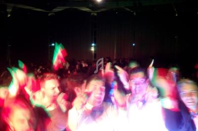 Personnes dansant dans une ambiance festive avec des lumières colorées, un drapeau visible en arrière-plan.