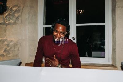 Un DJ souriant portant un casque audio, debout devant une table de mixage dans une pièce avec un mur en pierre.