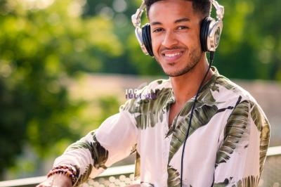 Un DJ souriant mixe de la musique en extérieur, entouré de verdure, avec un ordinateur portable à ses côtés.