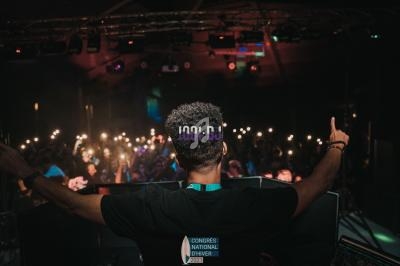 Un DJ de dos anime une foule dans une salle sombre, éclairée par des lumières et des téléphones allumés.