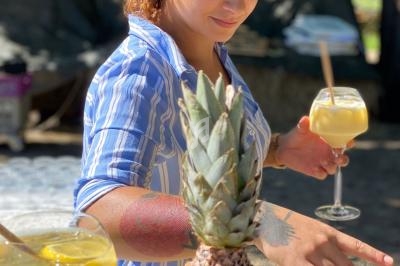 Une femme en chemise rayée prépare des boissons tropicales sur un comptoir extérieur, avec un ananas au premier plan.