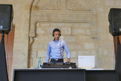 Un DJ en chemise bleue mixe de la musique devant un mur en pierre, entouré de deux enceintes.