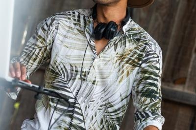 Un DJ souriant portant un chapeau et une chemise à motifs tropicaux mixe de la musique sur des platines en extérieur.