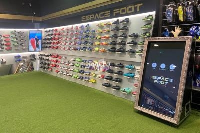 Mur d'exposition de chaussures de football colorées dans un magasin avec écran interactif et sol en gazon synthétique.