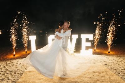 Un couple danse sur une plage illuminée par des lettres géantes ’LOVE’ et des feux d'artifice en arrière-plan.