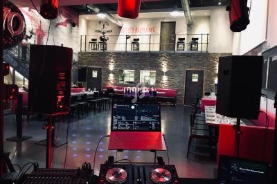 Vue depuis une cabine de DJ avec platines et ordinateur, dans un restaurant spacieux à l'éclairage tamisé.