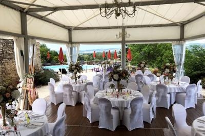 Salle de réception en plein air avec tables rondes décorées, chaises blanches et vue sur une terrasse avec parasols rouges.