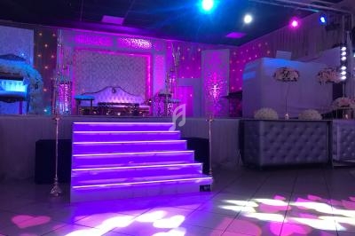 Scène décorée avec des lumières roses et violettes, escalier central illuminé, mobilier élégant et fleurs blanches.