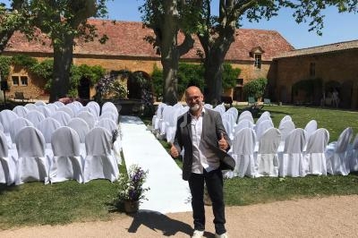 Un homme souriant pose devant une allée centrale bordée de chaises blanches dans un jardin, prêt pour un événement.