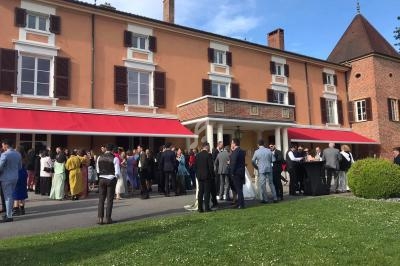 Groupe de personnes rassemblées devant un grand bâtiment aux murs orangés avec des stores rouges, par une journée…