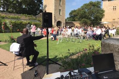 Un musicien joue devant un public assis lors d'une cérémonie en plein air dans un jardin avec des bâtiments en pierre.