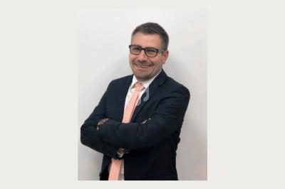 Homme en costume sombre avec une cravate rose, bras croisés, souriant devant un fond blanc uni.