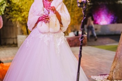 Une femme en robe de mariée blanche sourit devant un anneau lumineux, tenant un verre dans un cadre extérieur.