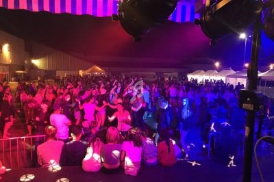 Foule dansant sous des éclairages colorés lors d'un événement nocturne en extérieur avec des stands et une scène.