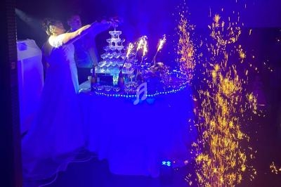 Une mariée verse du champagne dans une fontaine de verres illuminée, entourée de décorations et de gerbes d'étincelles.