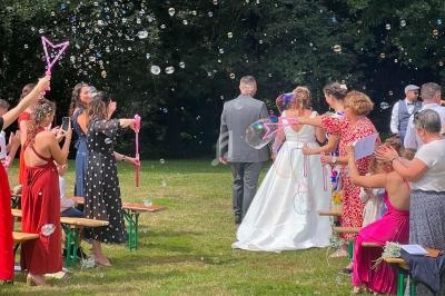 Des invités soufflent des bulles de savon autour d'un couple en tenue de mariage avançant sur une pelouse.