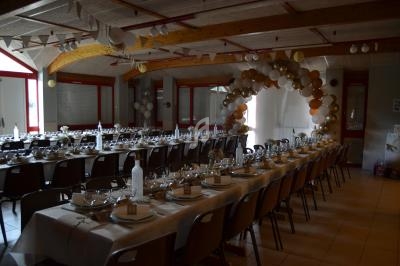 Salle décorée pour un événement avec longues tables dressées, vaisselle élégante et arche de ballons en arrière-plan.
