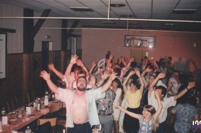 Des personnes dans une salle festive, levant les bras en l'air, entourées de tables avec des bouteilles et des verres.