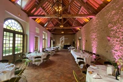 Salle de réception avec tables rondes dressées, chaises pliantes et éclairage chaleureux sous une charpente en bois.