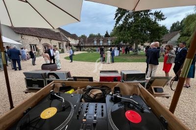 Vue depuis une table de mixage avec platines vinyles, donnant sur une réception en extérieur devant des bâtiments en pierre.