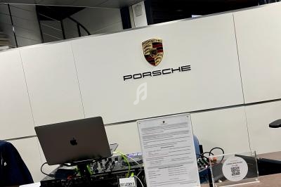 Bureau d'accueil avec logo Porsche sur un mur blanc, ordinateur et documents posés sur le comptoir en bois.