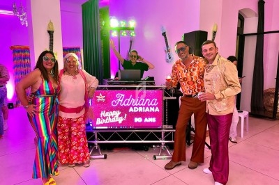 Des personnes en tenues colorées des années 70 posent devant un DJ et une pancarte lumineuse d'anniversaire.