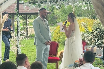 Une mariée en robe blanche prononce un discours face à son époux sous une tente lors d'une cérémonie en plein air.