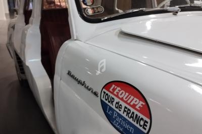 Voiture ancienne blanche avec autocollant ’L'Équipe Tour de France’, porte ouverte et vélos fixés sur le toit.