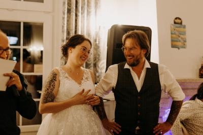 Un couple souriant, la mariée en robe blanche et le marié en gilet, dans une ambiance chaleureuse en intérieur.