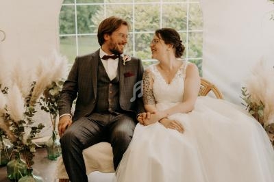 Un couple en tenue de mariage assis sur une chaise dans une tente décorée de plantes et de pampas.