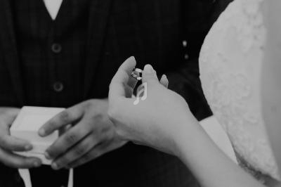 Une mariée tenant une alliance tandis qu'un marié en costume sombre se tient en arrière-plan.