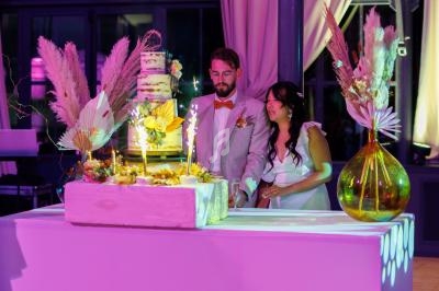 Un couple en tenue de mariage se tient près d'une table décorée avec un gâteau et des fleurs, dans une salle éclairée.