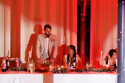 Un homme debout et deux femmes assises à une table décorée, dans une salle éclairée par une lumière rouge.