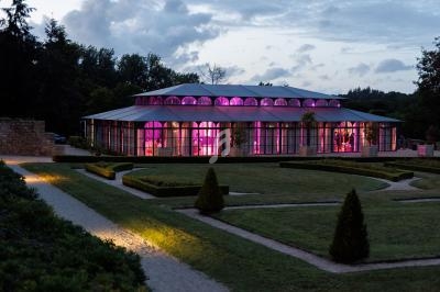Serre illuminée en rose au crépuscule, entourée de jardins symétriques et de chemins gravillonnés.