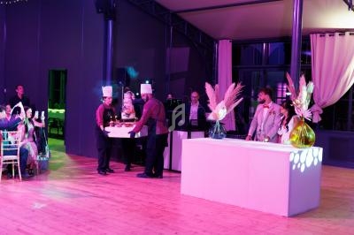 Des chefs apportent un gâteau de mariage décoré dans une salle de réception éclairée avec des invités en arrière-plan.